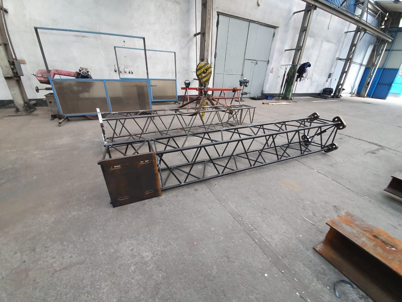 Konstrukcje metalowe kratownicowe, prawdopodobnie do banerów reklamowych, w hali produkcyjnej w Szczecinie. Widoczny surowy beton posadzki, brama w tle oraz elementy wyposażenia warsztatu.