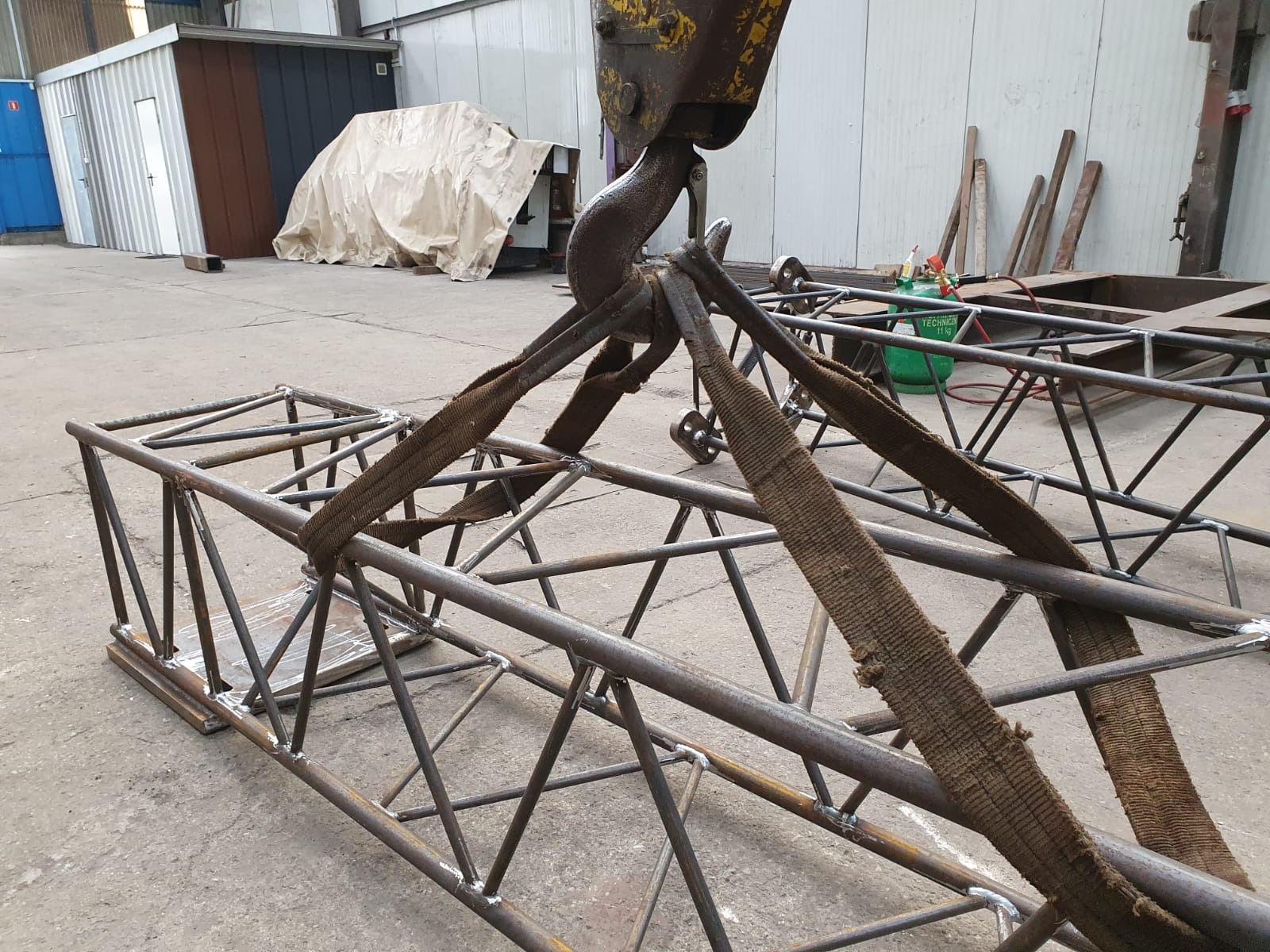 Metalowa konstrukcja kratowa, podnoszona za pomocą pasów i haka dźwigu w hali produkcyjnej, widoczne spawy i elementy montażowe.