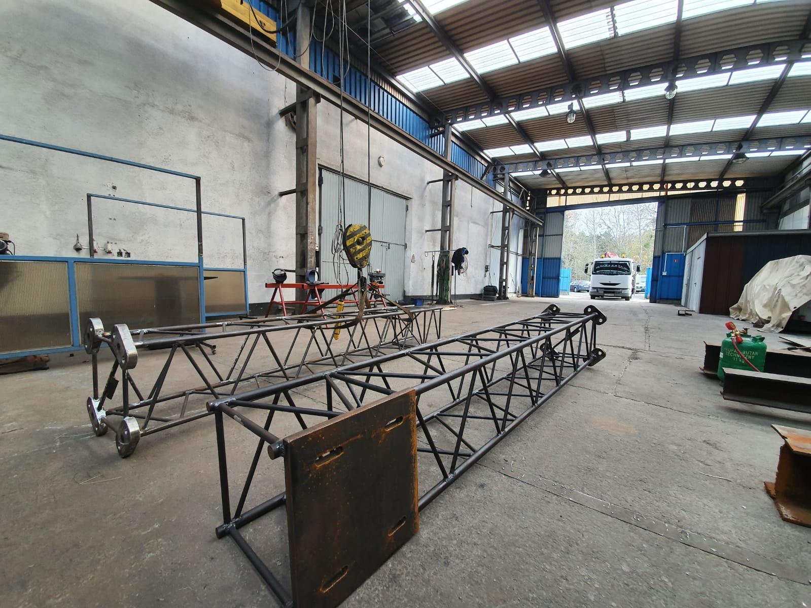 Dwie metalowe konstrukcje kratownicowe, prawdopodobnie do banerów reklamowych, leżą na posadzce hali produkcyjnej. Jedna konstrukcja z zamontowanymi kółkami transportowymi. W tle widoczny otwarty...