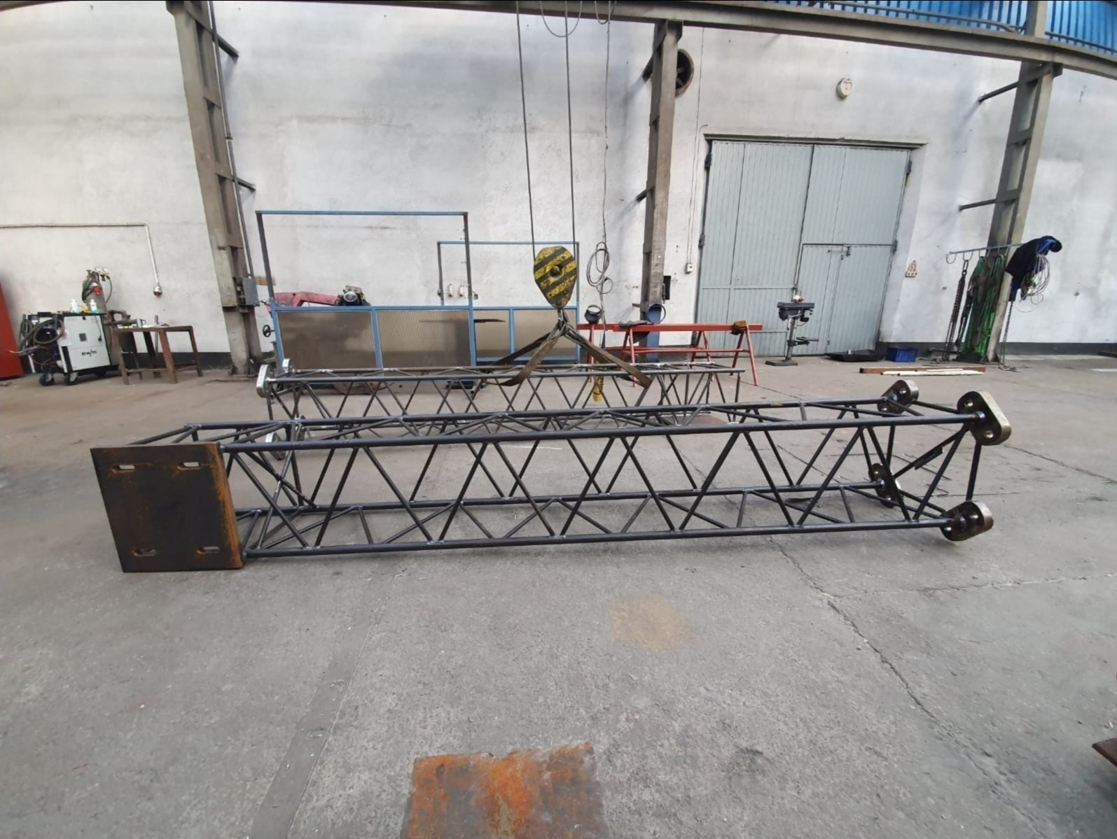 Metalowe konstrukcje kratownicowe, prawdopodobnie do banerów reklamowych, leżące na betonowej podłodze w hali produkcyjnej, widoczne spawarki i inne elementy wyposażenia warsztatu.