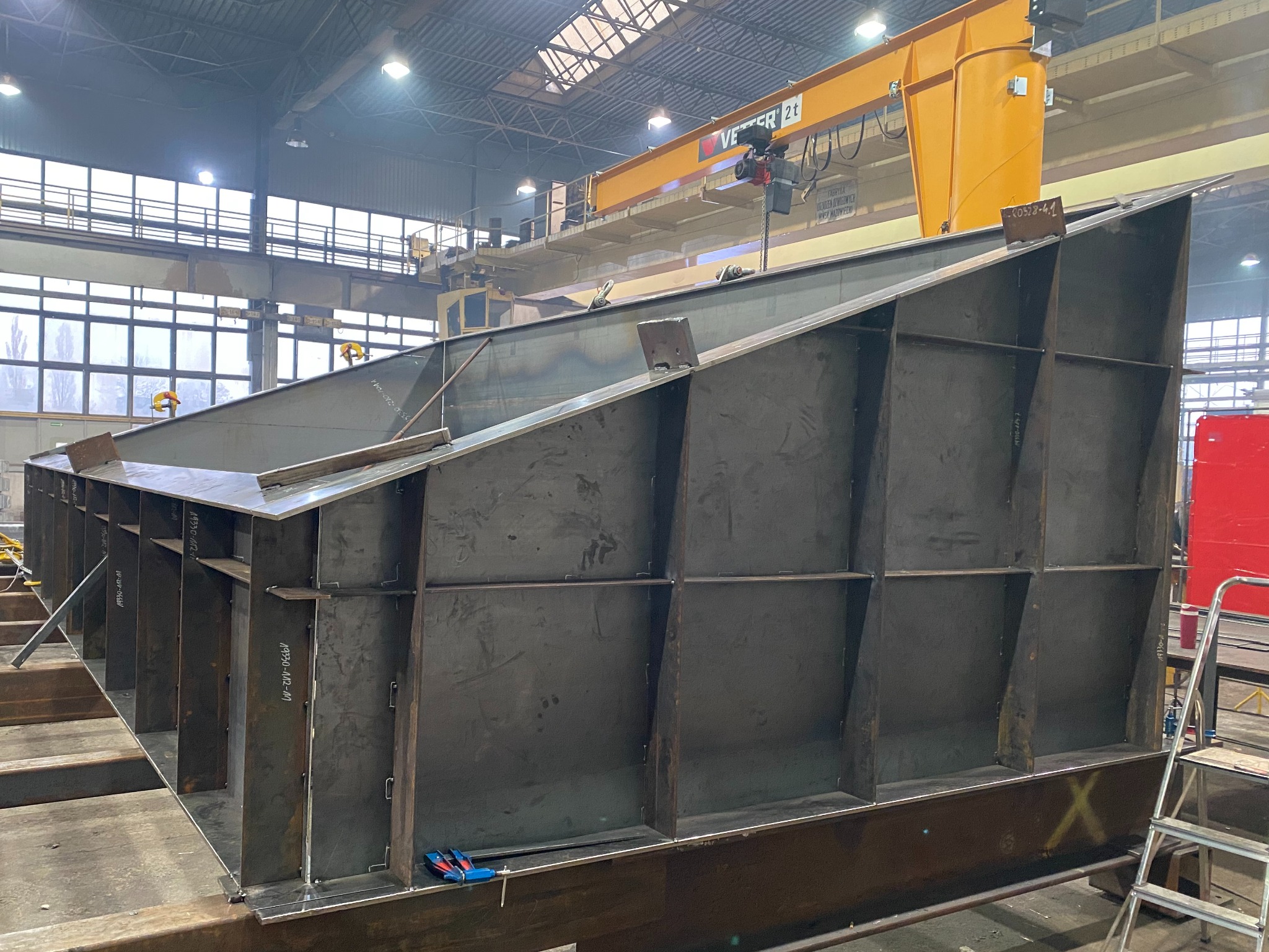 Konstrukcja stalowa z ukośnymi ścianami i wewnętrznymi wzmocnieniami, częściowo spawana, z żółtym suwnicą pomostową w tle hali przemysłowej.