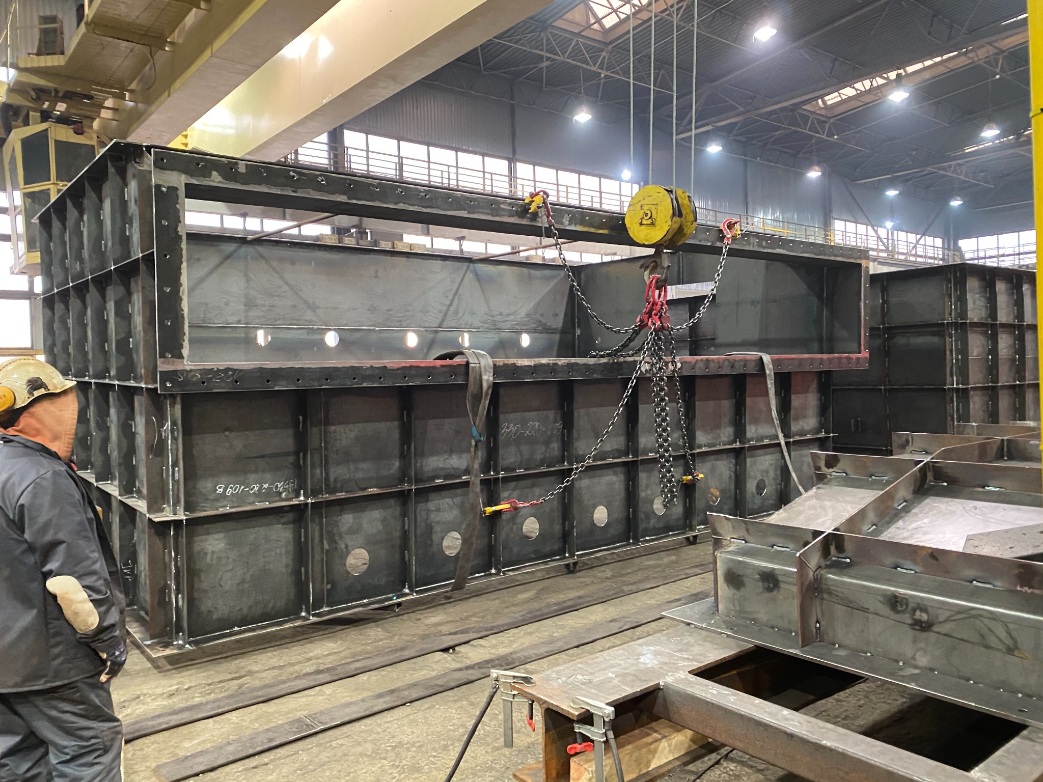 Wnętrze hali produkcyjnej z surową, spawaną konstrukcją stalową podnoszoną przez suwnicę linową; widoczny pracownik w kasku i roboczej odzieży.