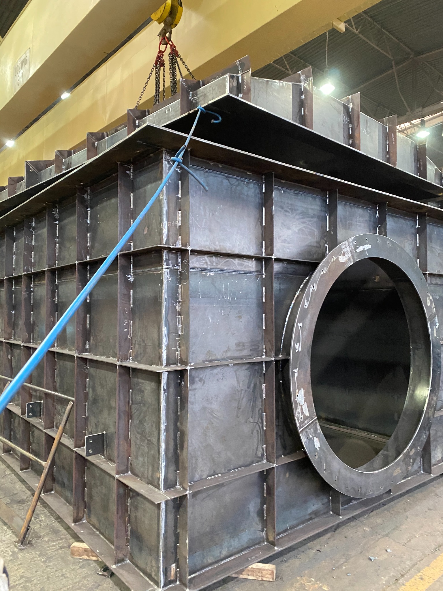 Konstrukcja metalowa spawana z otworem kołowym, podnoszona przez suwnicę z żółtym hakiem. Widoczne liczne spawy i wzmocnienia.