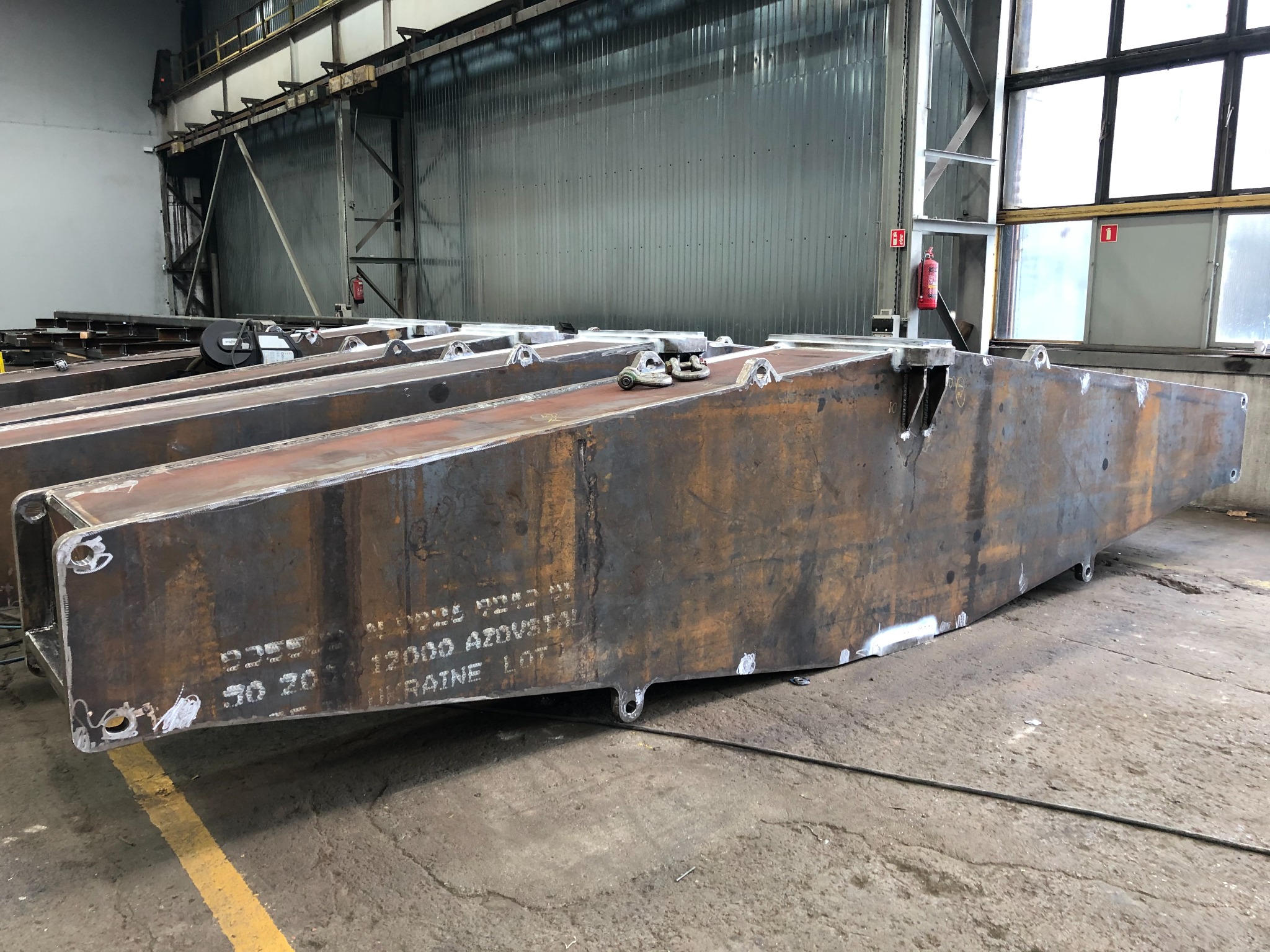 Surowe, metalowe elementy konstrukcyjne w hali produkcyjnej, z widocznymi śladami obróbki i spawania, leżące na betonowej podłodze, z napisami technicznymi na powierzchni.