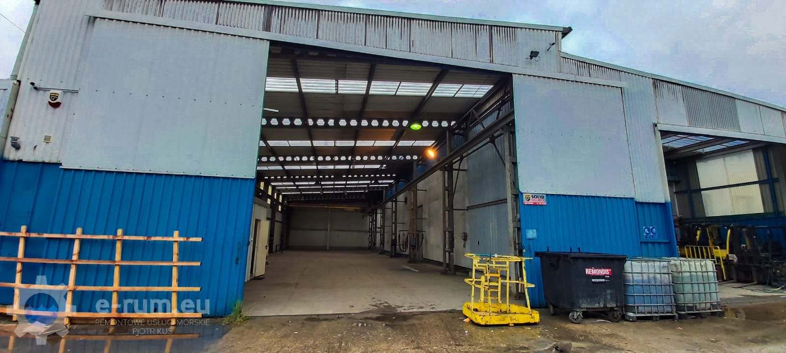 Szeroka brama wjazdowa do hali przemysłowej z szarej blachy falistej z niebieskim dołem, widoczne wnętrze hali z suwnicą, na zewnątrz stoją kontenery i podnośnik.