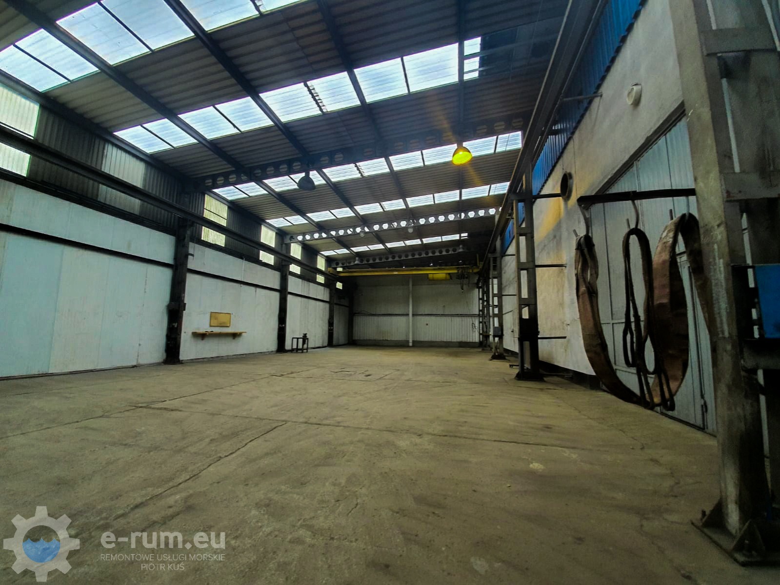 Wnętrze hali produkcyjnej z suwnicą pod sufitem, oświetlone przez świetliki dachowe i pojedynczą lampę, z widocznymi elementami konstrukcji stalowej i zawieszonymi pasami transportowymi.