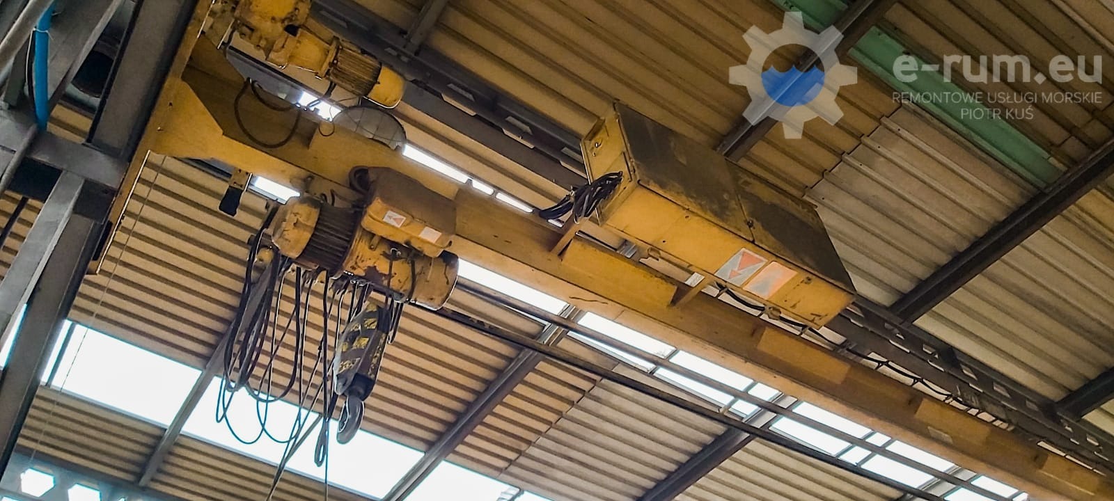 Żółty suwnicowy dźwig z silnikami i hakiem podwieszony pod metalowym dachem hali przemysłowej, widok z dołu.