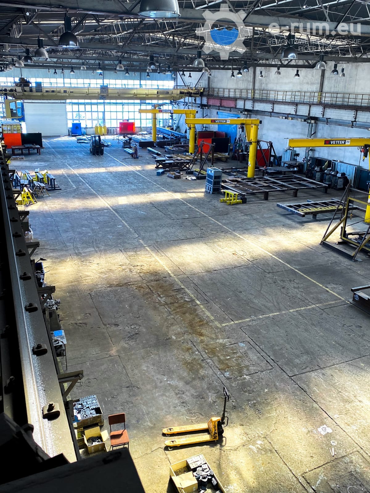 Wnętrze hali produkcyjnej z suwnicami stanowiskowymi Vetter 2T, widok z góry na betonową posadzkę z żółtymi liniami, w tle widoczny wózek widłowy i elementy konstrukcji stalowych.