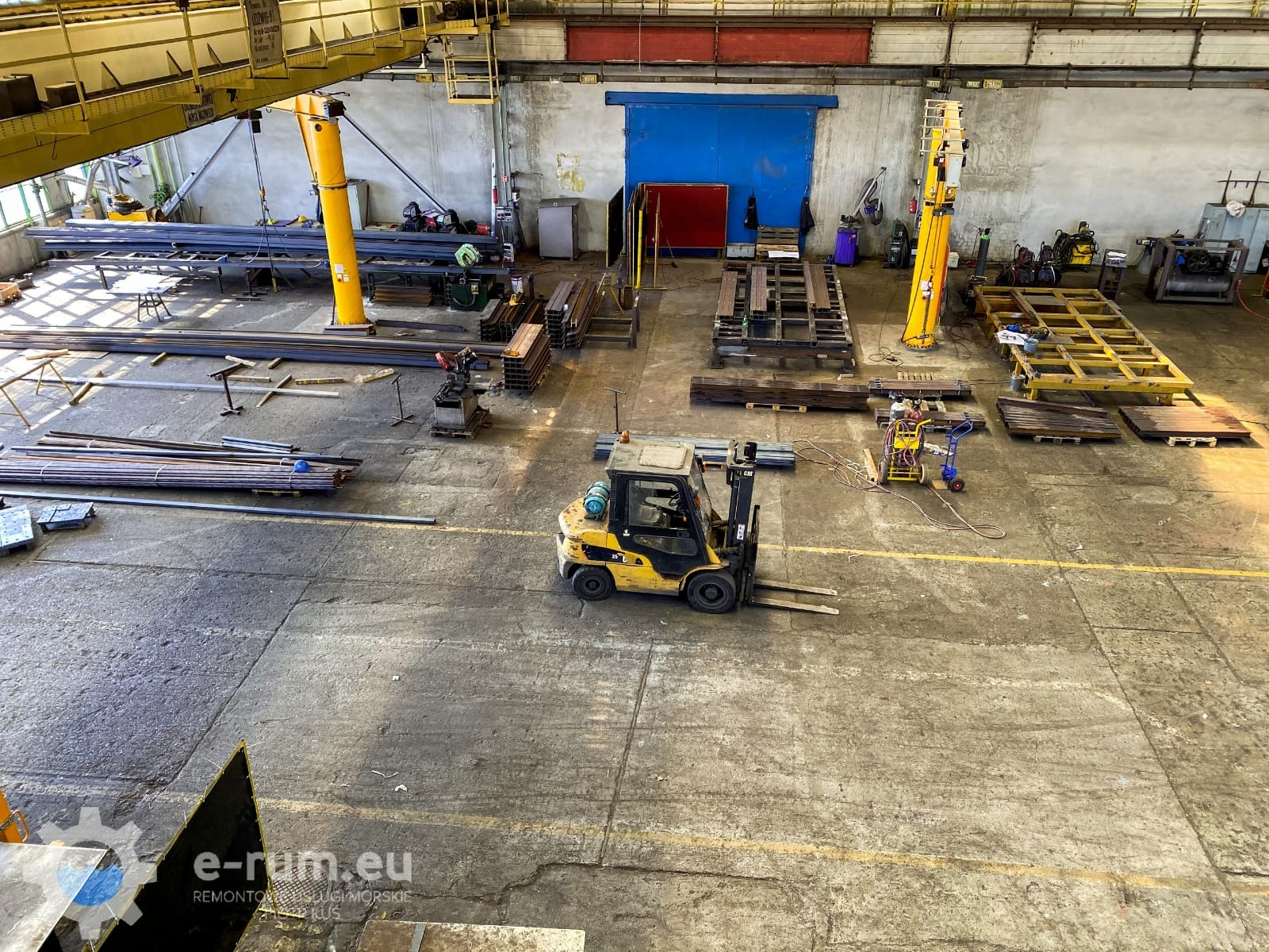 Wnętrze hali produkcyjnej z surowymi elementami metalowymi, wózkiem widłowym i urządzeniami spawalniczymi, widok z góry.