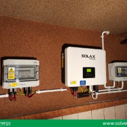Rozdzielnica PV-DC, inwerter Solax Power X3 i dodatkowa skrzynka zabezpieczeń połączone przewodami w instalacji wewnętrznej z brązowym tłem, widoczne elementy instalacji elektrycznej.