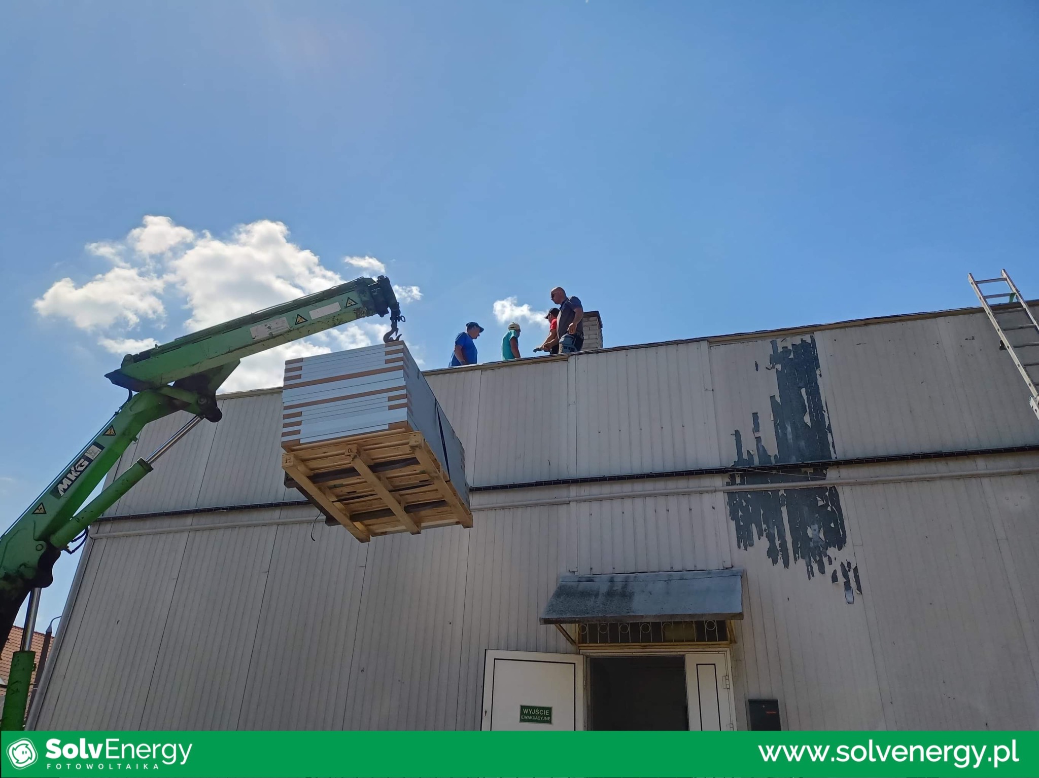 Montaż paneli na dachu budynku przemysłowego za pomocą dźwigu, ekipa monterów na dachu, błękitne niebo z chmurami w tle.