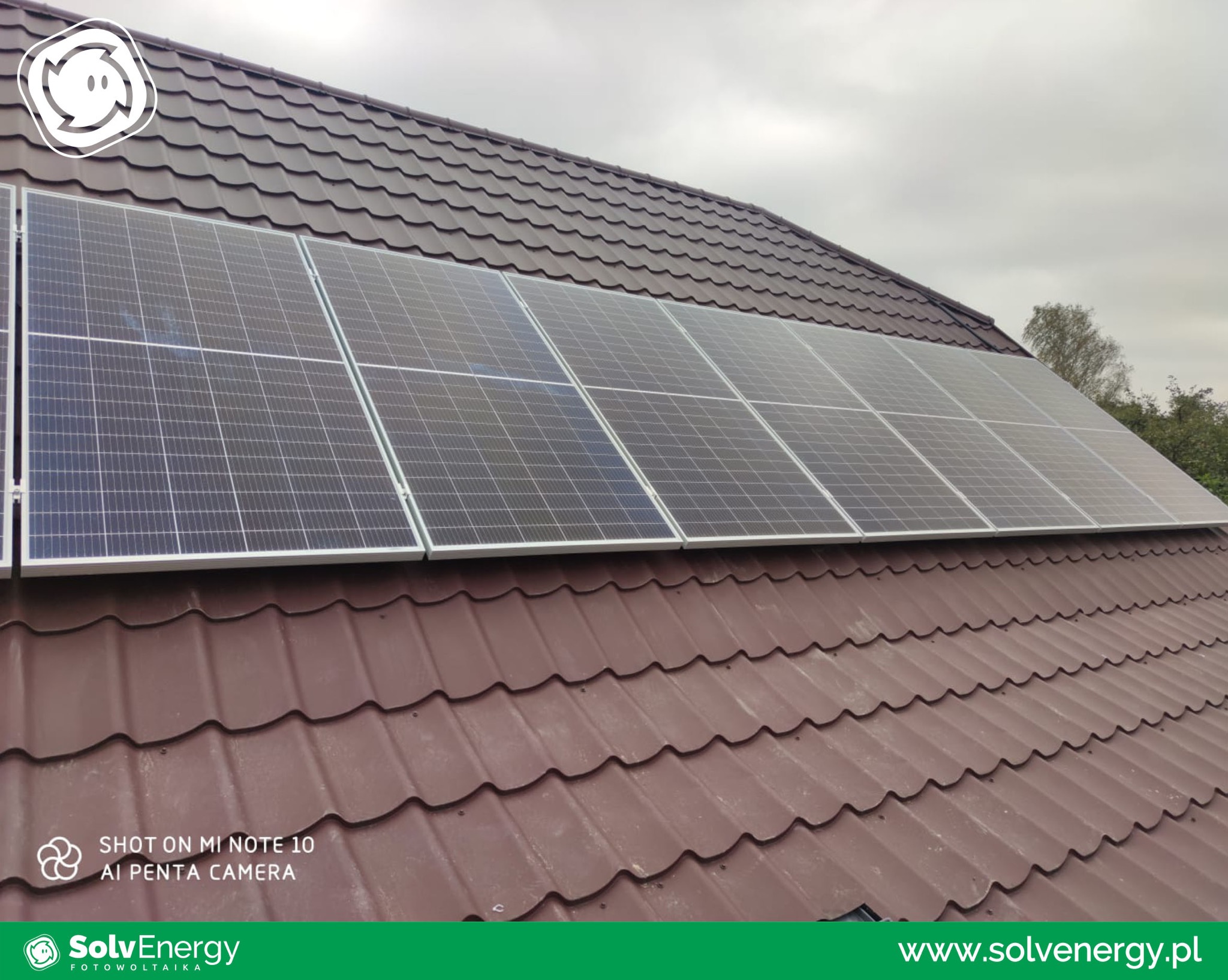 Panele słoneczne zamontowane na brązowym dachu z blachodachówki, pochmurne niebo w tle.