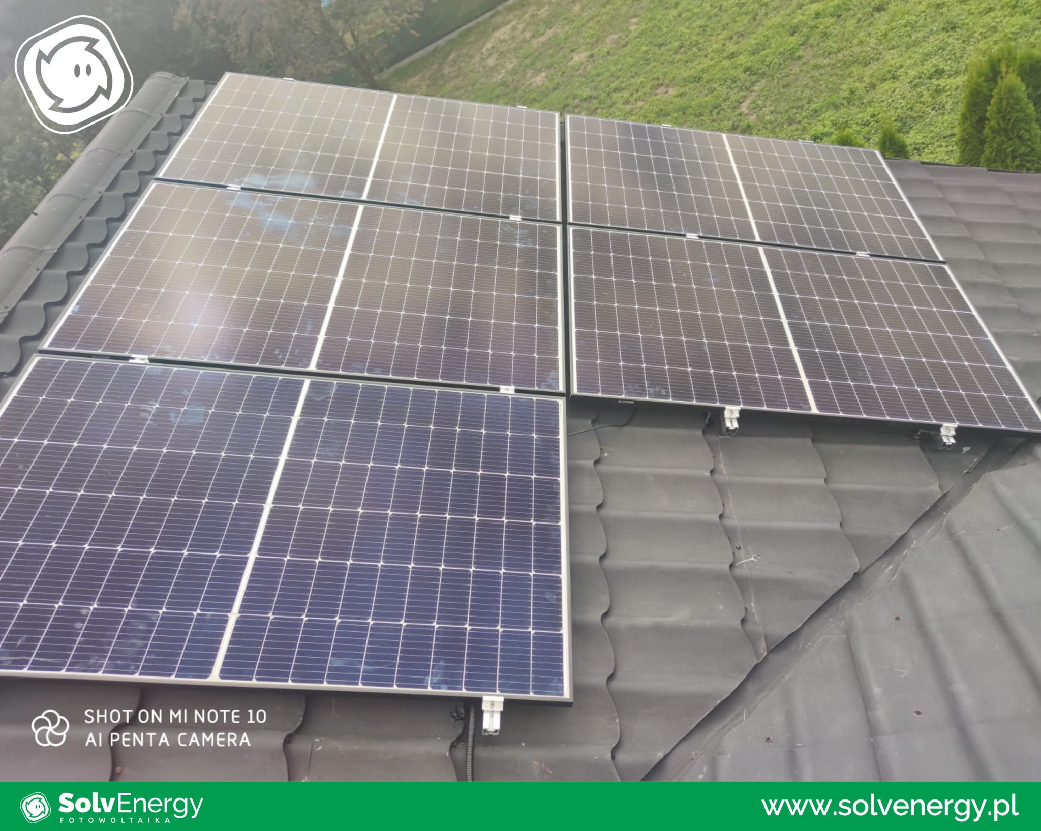 Panel fotowoltaiczny na ciemnej, falistej dachówce, widoczne mocowania i kable, zieleń w tle, logo firmy w dolnej części kadru.
