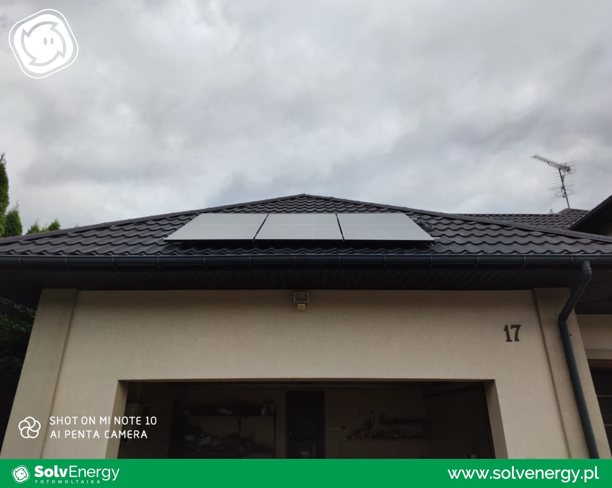 Trzy panele słoneczne na dachu domu z ciemną dachówką, widoczne rynny i numer domu '17', pochmurne niebo w tle.