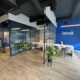 Nowoczesne biuro z przeszklonymi ścianami, białymi biurkami i szarymi krzesłami, logo 'lendi' na niebieskiej ścianie, rośliny doniczkowe w minimalistycznych stojakach.