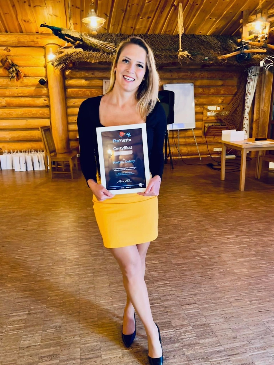 Uśmiechnięta blondynka w czarnej bluzce i żółtej spódnicy prezentuje certyfikat FinFiesta w drewnianym wnętrzu z dekoracjami.