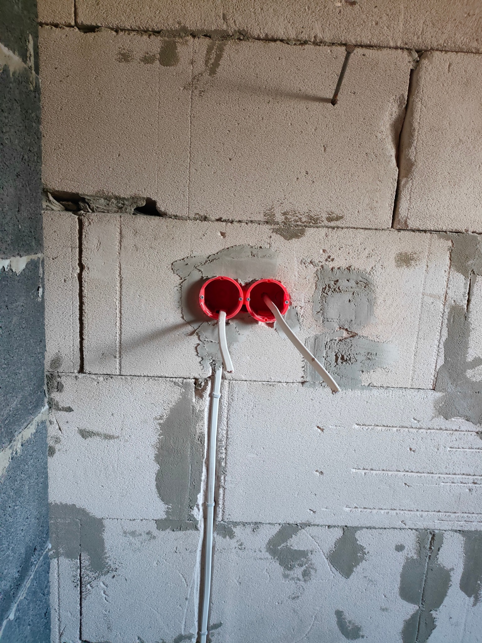 Dwa czerwone puszki elektryczne zamontowane w surowej ścianie z betonu komórkowego, z wystającymi białymi przewodami elektrycznymi.
