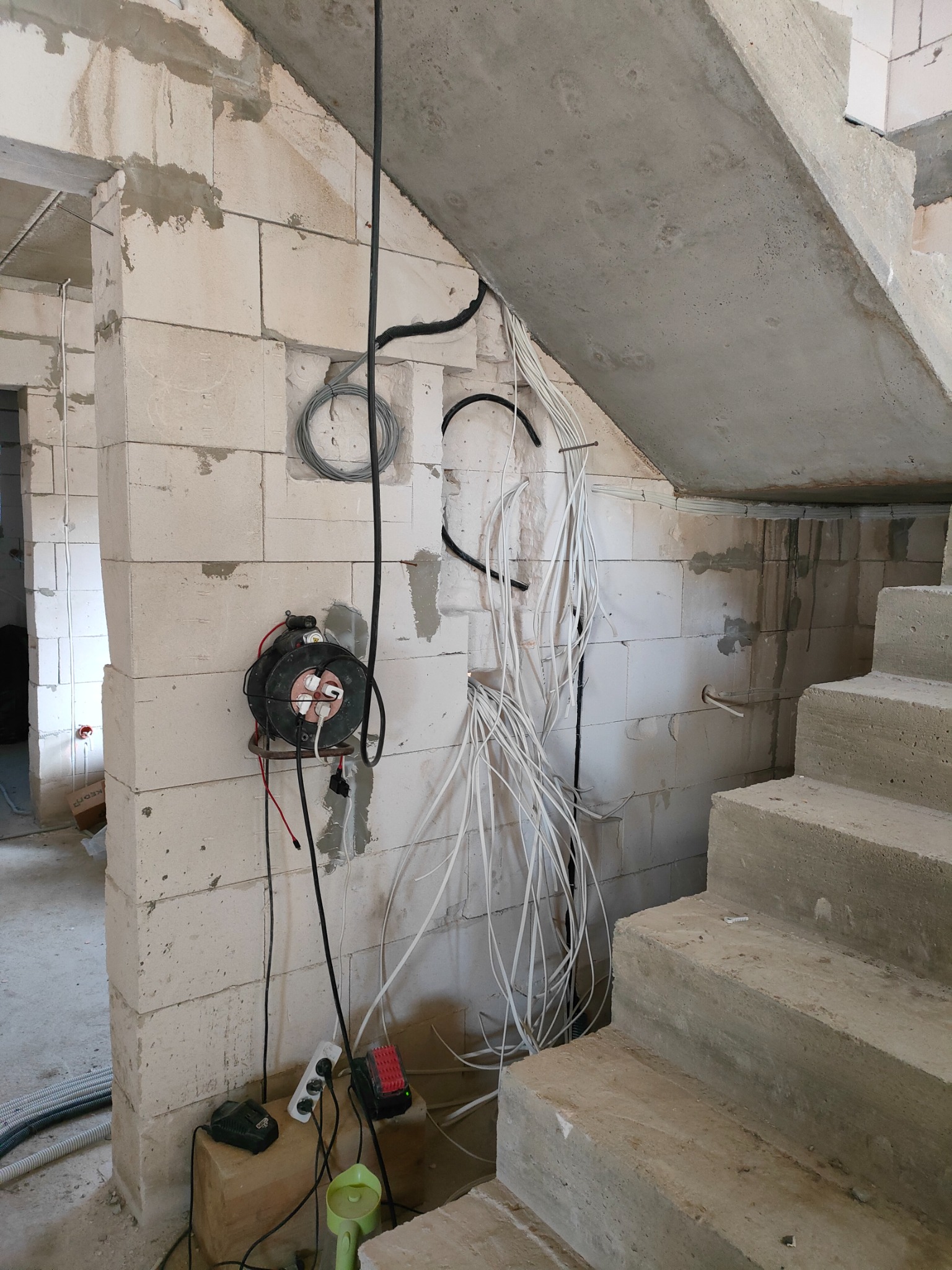 Surowy stan budynku w trakcie prac: widoczne przewody elektryczne wystające ze ściany z bloczków, rozdzielnica na ścianie, betonowe schody w tle.