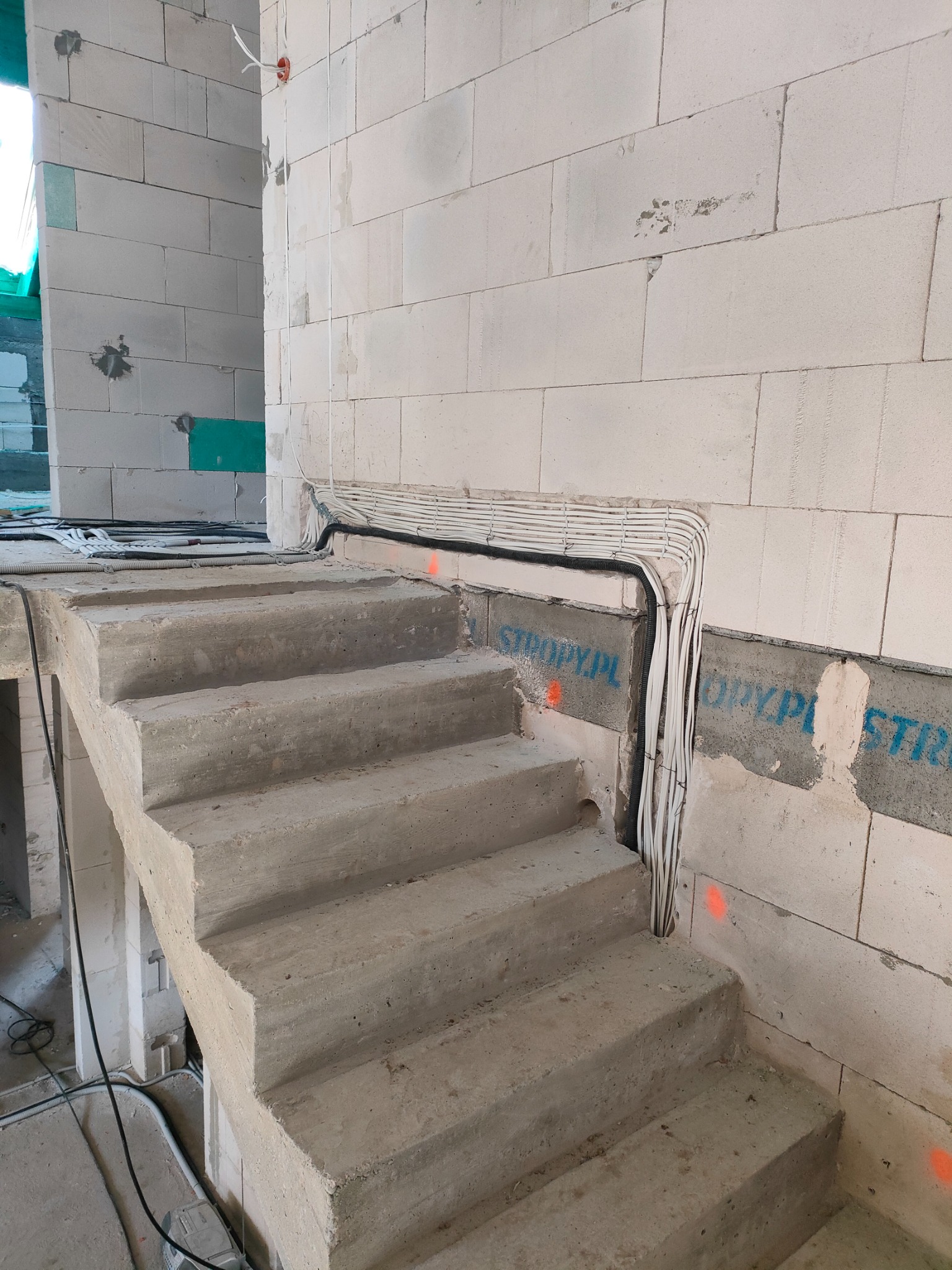 Surowe betonowe schody w trakcie budowy, z widocznymi przewodami elektrycznymi prowadzonymi wzdłuż ściany z bloczków, pomalowane napisy na ścianie, widoczne ślady prac budowlanych.