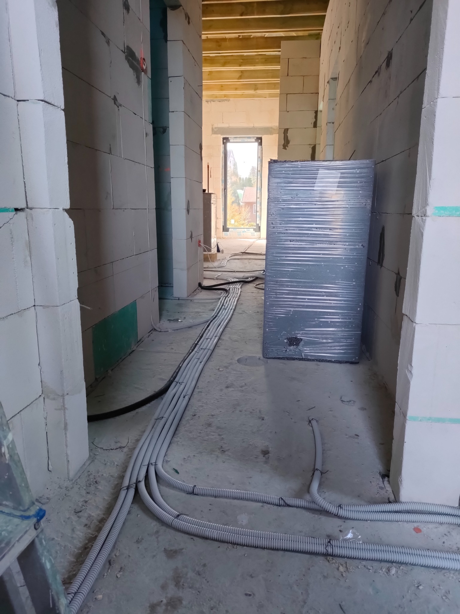 Wnętrze w trakcie budowy z widocznymi przewodami elektrycznymi ułożonymi na podłodze, ściany z bloczków betonowych, w głębi okno z widokiem na ogród.