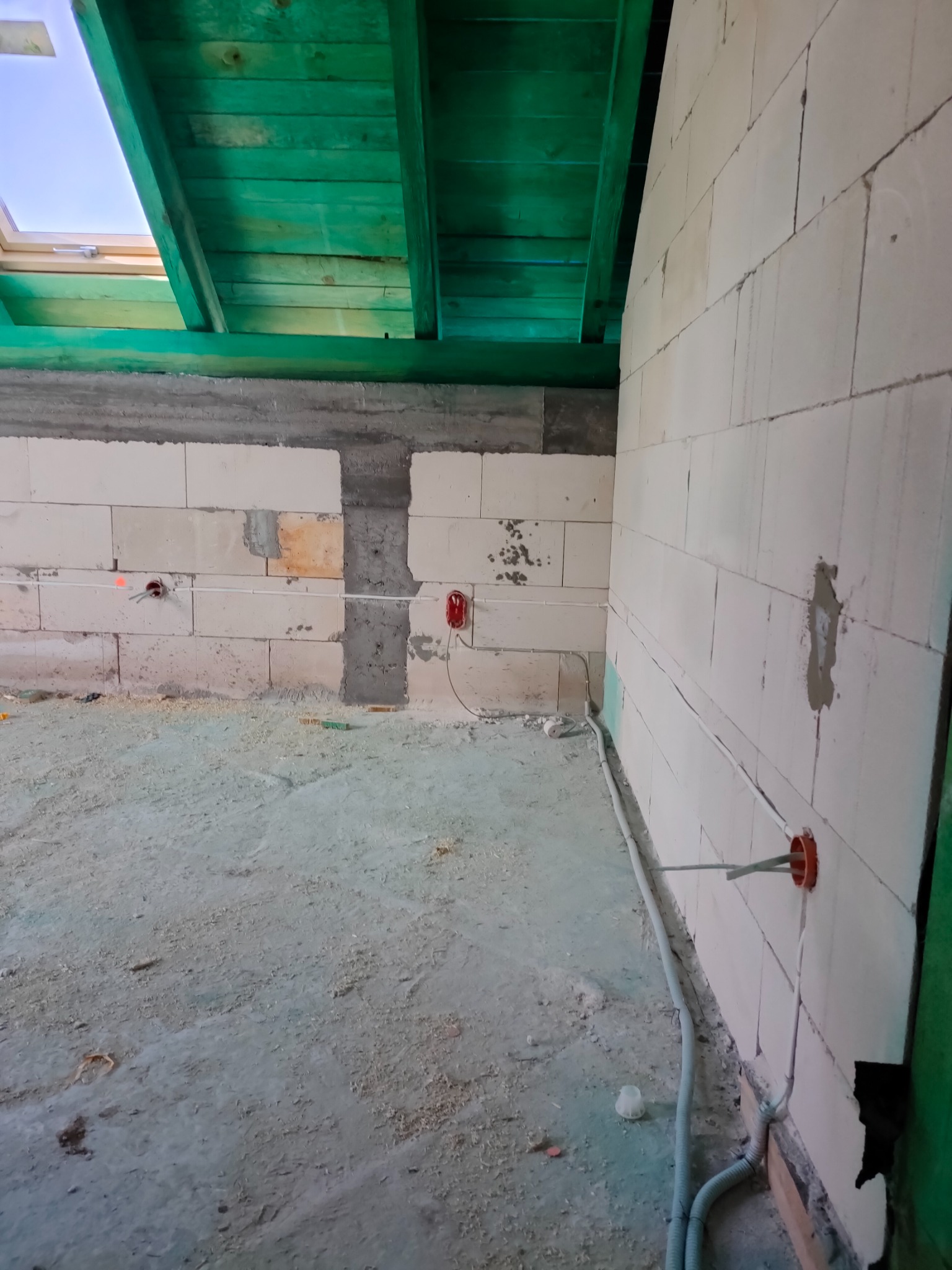 Surowy stan wnętrza z pustaków, widoczne kable elektryczne wystające ze ścian, puszki pod gniazdka, drewniana konstrukcja dachu pomalowana na zielono.