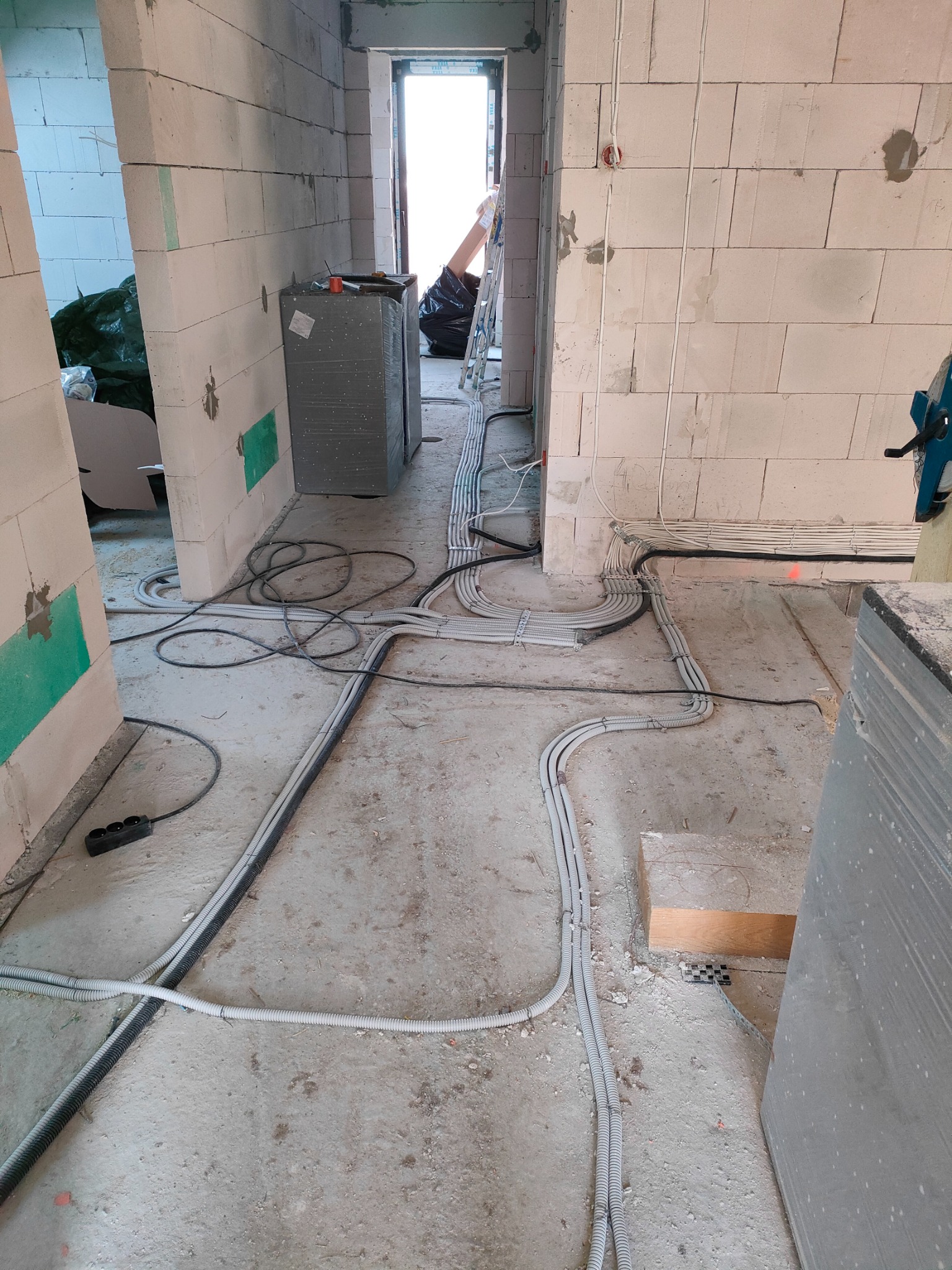 Instalacja elektryczna w surowym wnętrzu: kable w peszlach prowadzone po podłodze, ściany z betonu komórkowego w trakcie budowy, widoczny przedłużacz i drabina w tle.