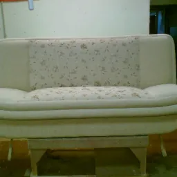 Odnowiona sofa w jasnej tapicerce z kwiatowym wzorem, umieszczona na drewnianym podeście w warsztacie.