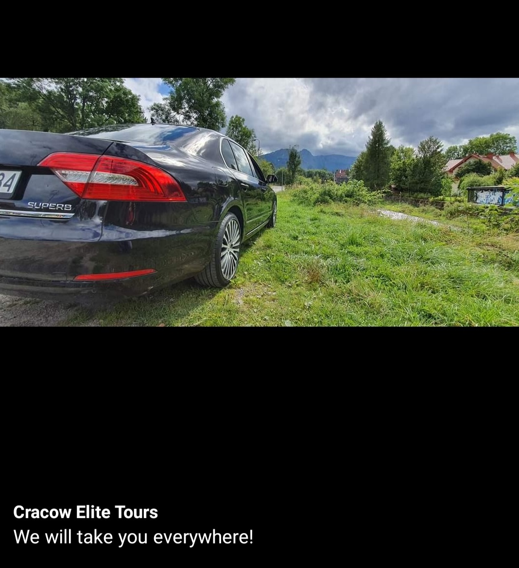 Czarny samochód osobowy marki Skoda Superb zaparkowany na trawiastym poboczu drogi, z widokiem na góry w tle, tablica rejestracyjna częściowo zasłonięta, napis 'Superb' widoczny na klapie bagażnika.