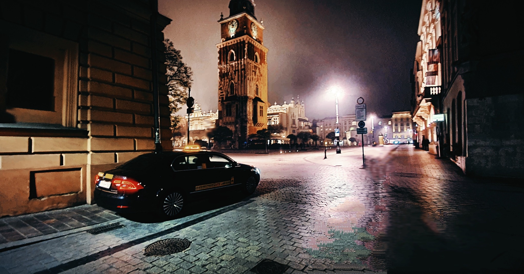 Czarna taksówka wysokiej klasy zaparkowana nocą na brukowanej ulicy w Krakowie, w tle Rynek Główny z Sukiennicami i Wieżą Ratuszową.