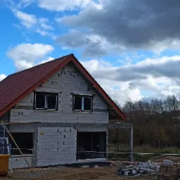 Surowy stan budynku z czerwonym dachem na tle pochmurnego nieba, widoczne rusztowania i teren budowy.