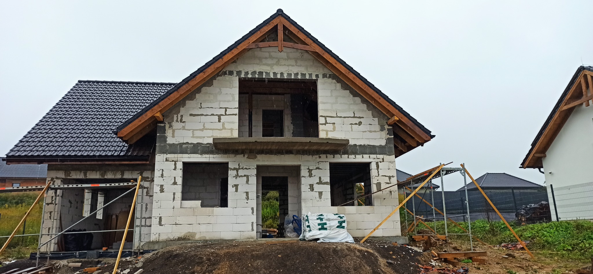 Surowy stan domu w trakcie budowy z widoczną konstrukcją dachu i częściowo położonymi dachówkami, ściany z białego bloczka, drewniana więźba dachowa i widoczne rusztowania.