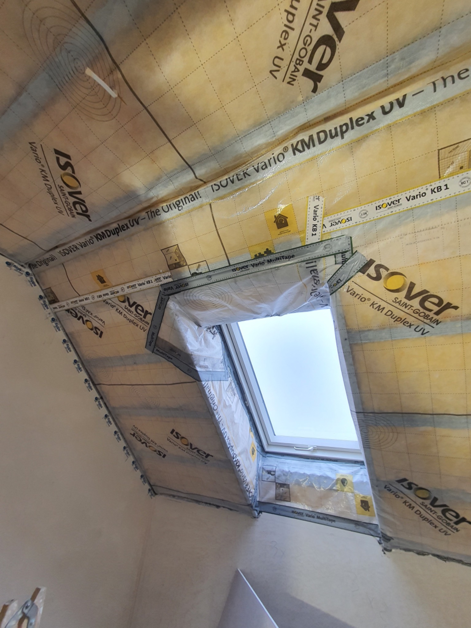 Szczegółowe ujęcie izolacji poddasza wokół okna dachowego, folia paroizolacyjna Isover Vario KM Duplex UV z widocznymi oznaczeniami i taśmami uszczelniającymi, w trakcie montażu.