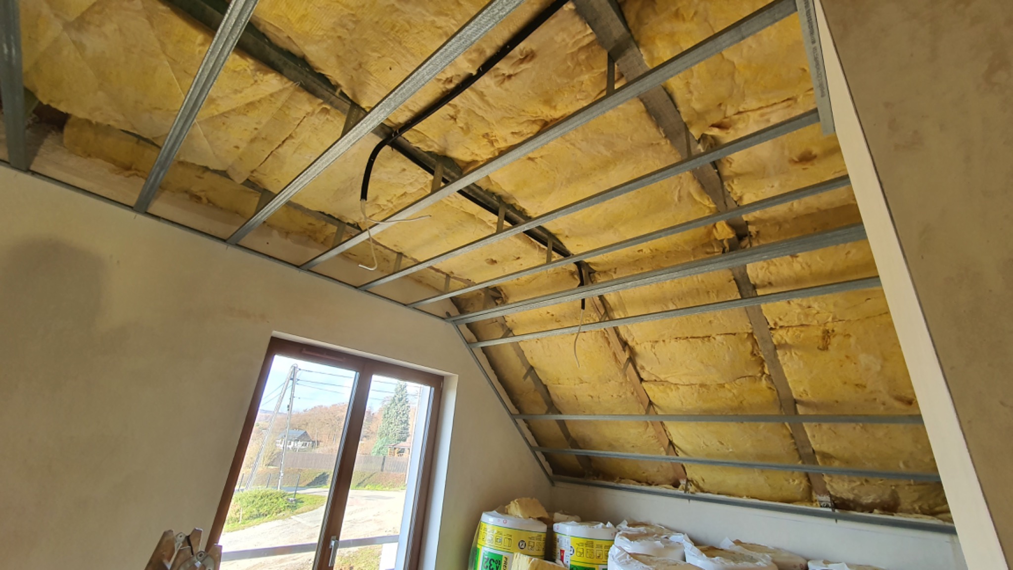 Poddasze w trakcie ocieplania wełną mineralną, widoczna metalowa konstrukcja sufitu podwieszanego, okno z widokiem na zewnątrz i worki z wełną u dołu kadru.