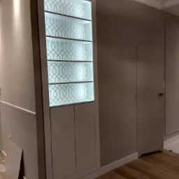 Wbudowana szafa z podświetlanymi półkami i dekoracyjnym wzorem na tylnej ściance, dopasowana kolorystycznie do ścian w odcieniach szarości i drewnianej podłogi.