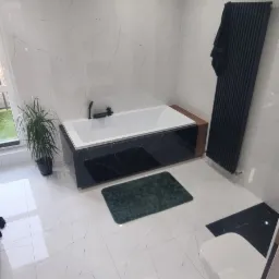 Nowoczesna łazienka z białą marmurową glazurą, czarną obudową wanny i grzejnikiem drabinkowym, uzupełniona zielonym dywanikiem i rośliną doniczkową przy oknie.