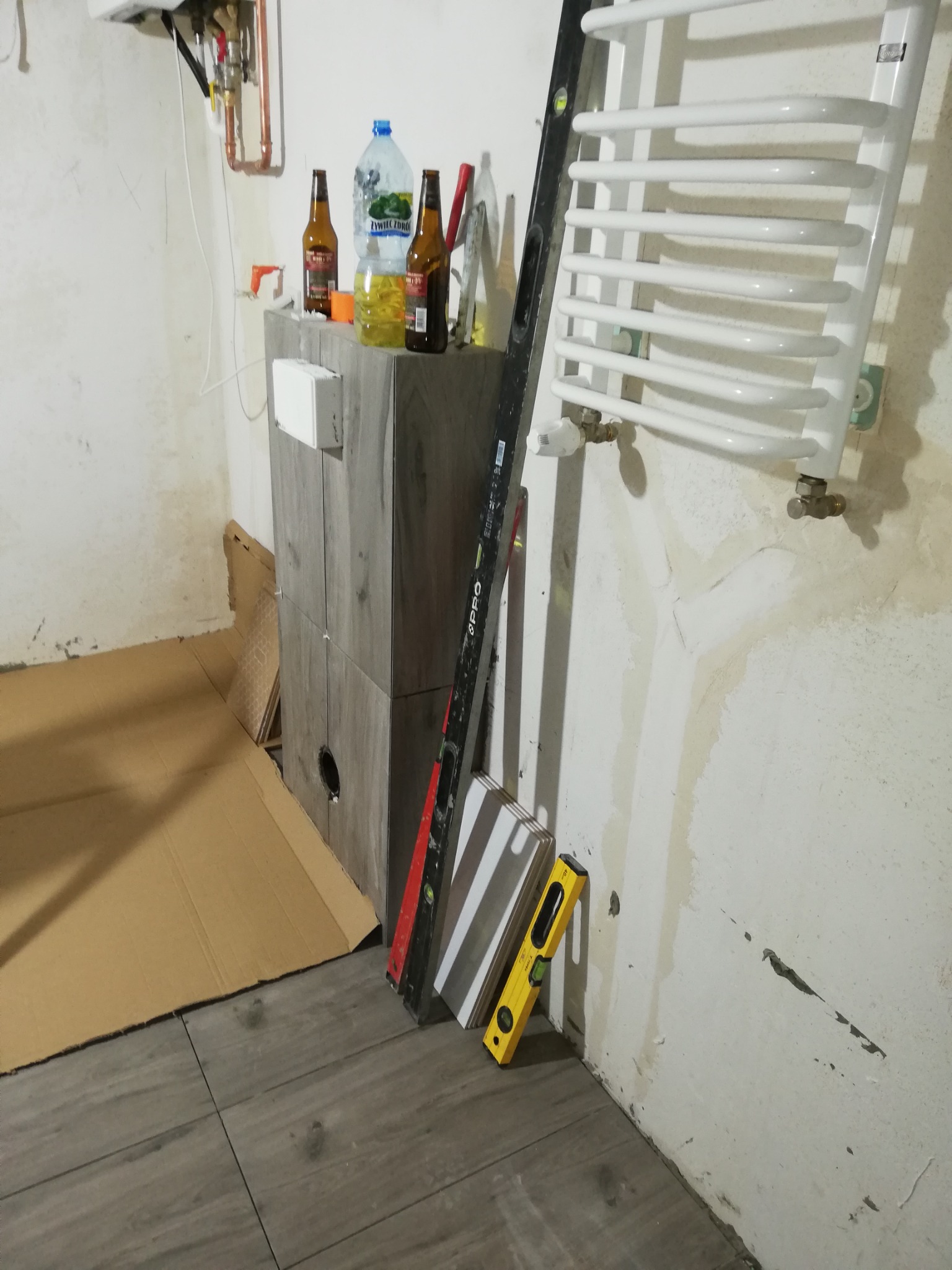 Fragment łazienki w trakcie remontu: ułożona szara podłoga z płytek imitujących drewno, przy ścianie oparty grzejnik drabinkowy, poziomice, obudowa rury zasilającej piec, na obudowie butelki...