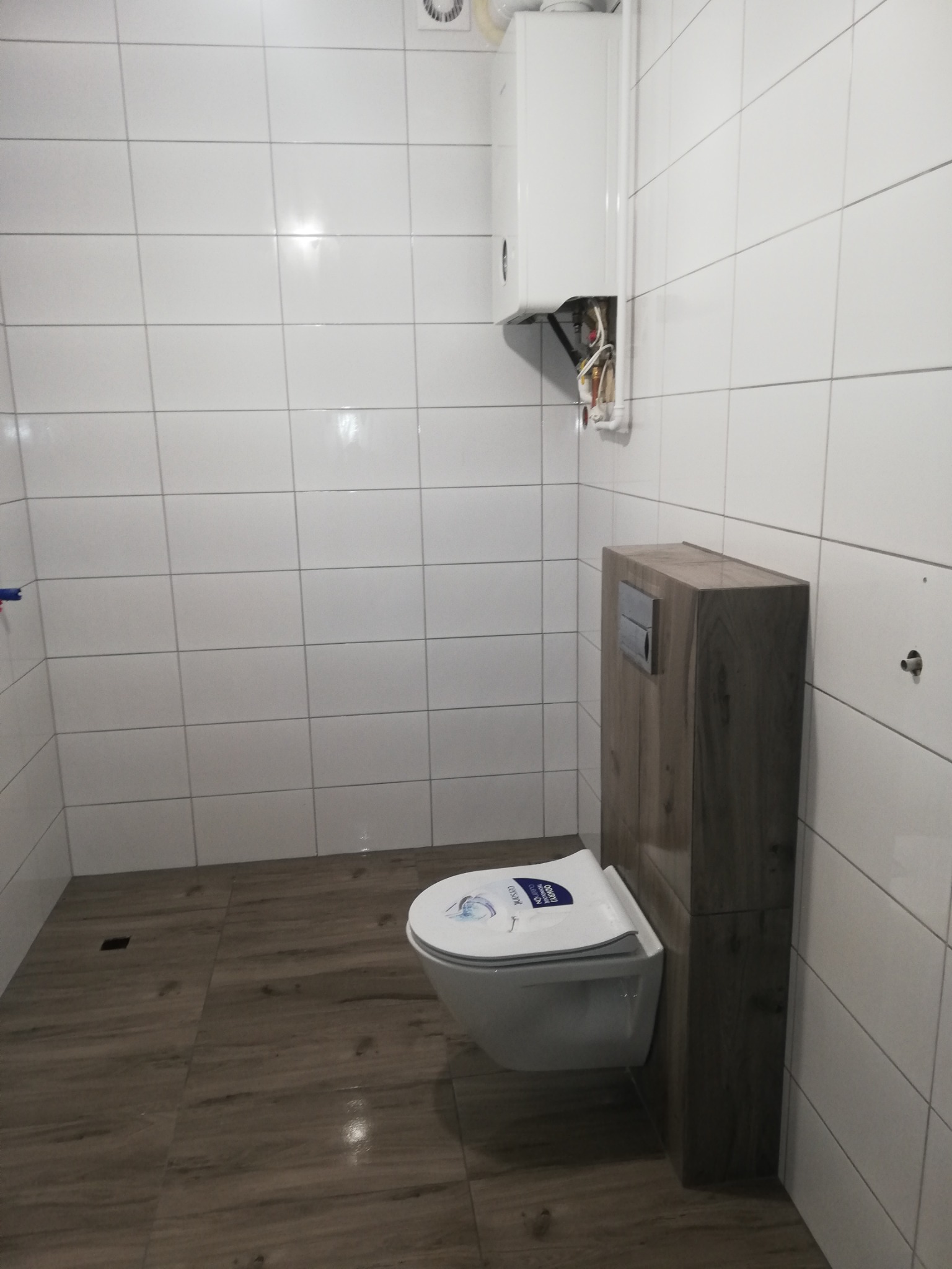 Nowa łazienka z podwieszaną toaletą z drewnianą obudową, białymi płytkami ściennymi i brązowymi płytkami podłogowymi imitującymi drewno, oraz wiszącym piecem gazowym z widocznymi przewodami.