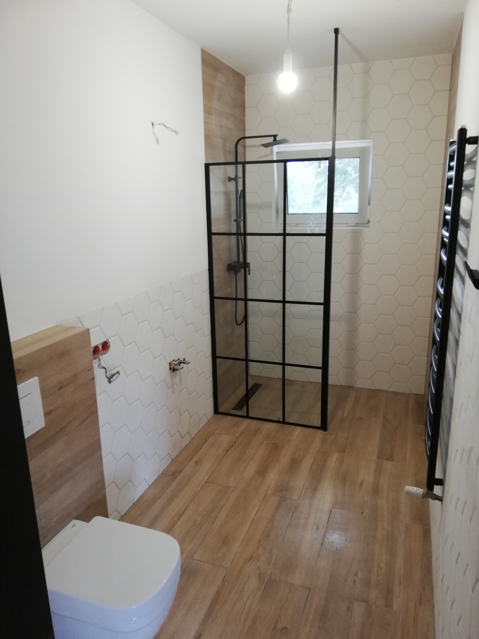 Wnętrze łazienki w trakcie wykańczania, widoczna kabina prysznicowa z czarną ramą, heksagonalne płytki na ścianach, drewnopodobne płytki na podłodze, oraz instalacja elektryczna i hydrauliczna...