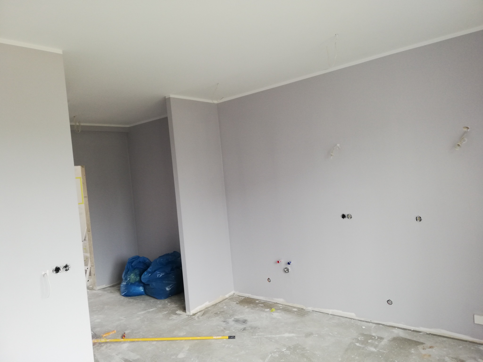 Szare ściany w trakcie remontu mieszkania, widoczne puszki elektryczne, rury instalacji wodnej, niebieskie worki z odpadami budowlanymi i żółty wałek malarski na podłodze.