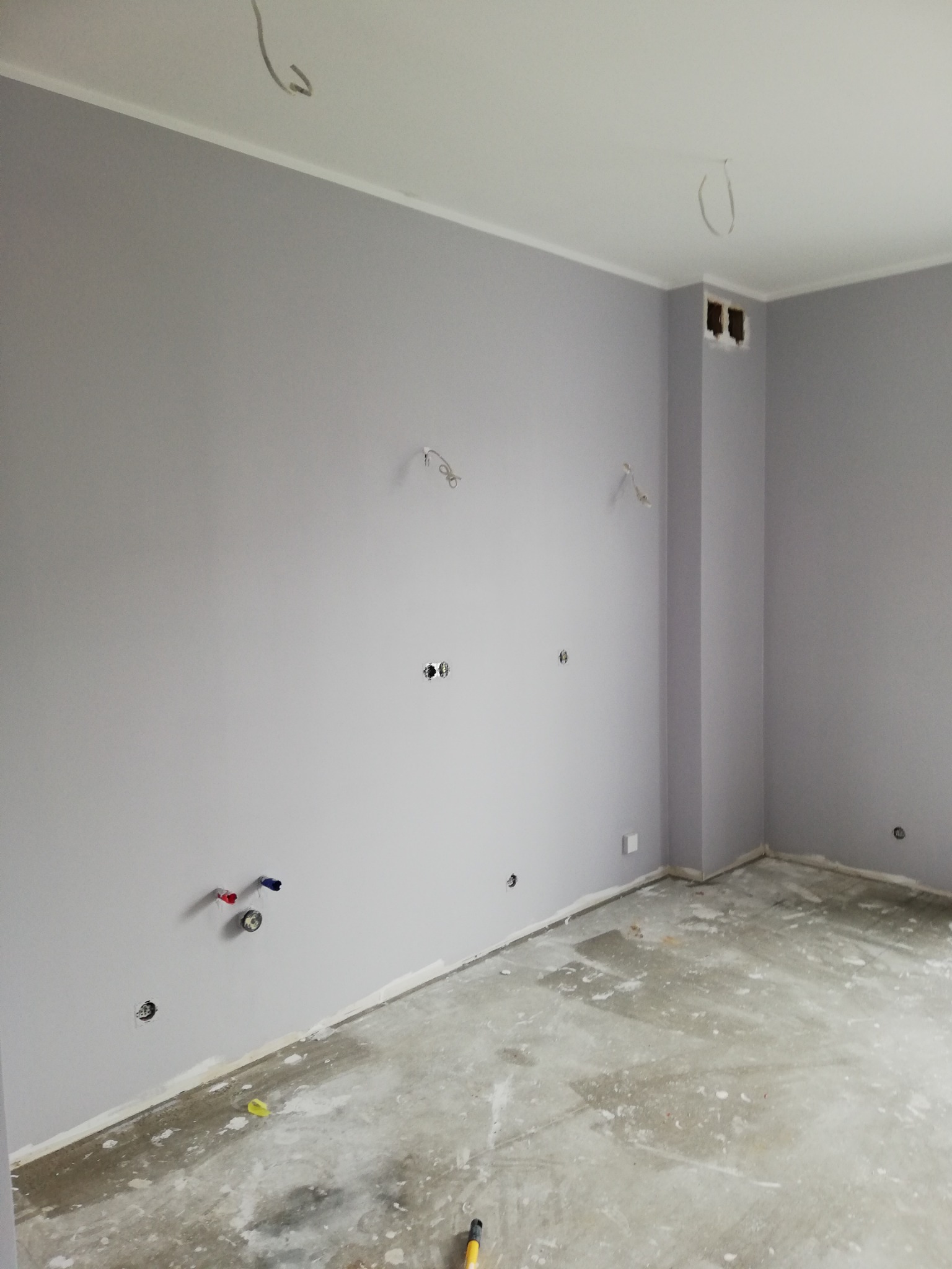 Pokój po malowaniu ścian na szaro, widoczne wyprowadzone instalacje elektryczne i wodne, surowa podłoga przygotowana do dalszych prac wykończeniowych.