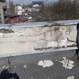 Zniszczona attyka na dachu budynku z widocznymi pęknięciami i odpadającym tynkiem, z fragmentami gruzu na pokryciu dachowym.