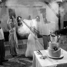 IvanTech Michał Iwanisik - Czarno-białe zdjęcie z wesela: para młoda i goście tańczą przy torcie weselnym z fontannami iskier, nagłośnienie i oświetlenie w tle.