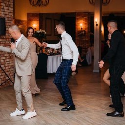 IvanTech Michał Iwanisik - Goście weselni tańczą na parkiecie, mężczyzna w beżowym garniturze śpiewa do mikrofonu, w tle kolumny obłożone cegłą i dekoracyjne żarówki.