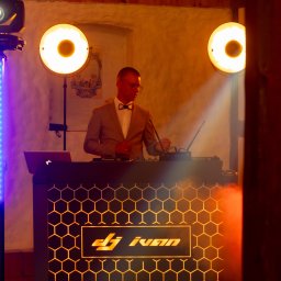 IvanTech Michał Iwanisik - DJ Ivan w trakcie występu, oświetlenie sceniczne z reflektorami i listwą LED, konsoleta z logo na tle ceglanej ściany.