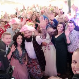 IvanTech Michał Iwanisik - Rozbawieni goście weselni w kolorowych perukach i eleganckich strojach, unoszący ręce w geście radości, z widocznym mikrofonem sugerującym obecność zespołu muzycznego lub wodzireja.