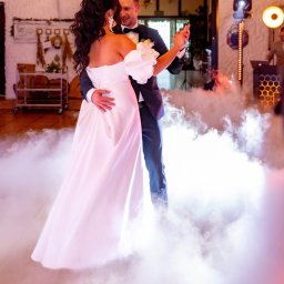 IvanTech Michał Iwanisik - Para młoda tańczy pierwszy taniec w otoczeniu sztucznego dymu, widoczne oświetlenie sceniczne i DJ za konsoletą.