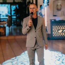 IvanTech Michał Iwanisik - Mężczyzna w okularach i szarym garniturze w kratę trzyma mikrofon, przemawiając na tle sceny z oświetleniem LED.
