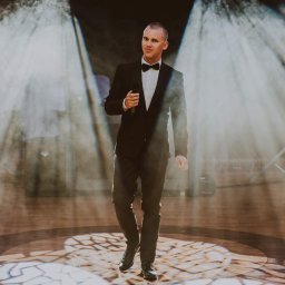 IvanTech Michał Iwanisik - Elegancko ubrany mężczyzna w muszce, trzymający mikrofon, idzie po scenie oświetlonej reflektorami.