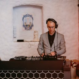 IvanTech Michał Iwanisik - DJ w szarej marynarce w kratę, skupiony na mikserze Pioneer DJ, słuchawki na uszach, pomarańczowe światło w tle, rustykalne wnętrze z białymi ścianami i dekoracjami.