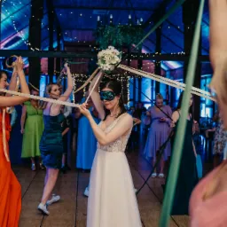 Panna młoda z zasłoniętymi oczami, trzymająca bukiet, uczestniczy w tradycyjnej zabawie weselnej z wstążkami, otoczona gośćmi w sali z drewnianą konstrukcją i dekoracyjnym oświetleniem.